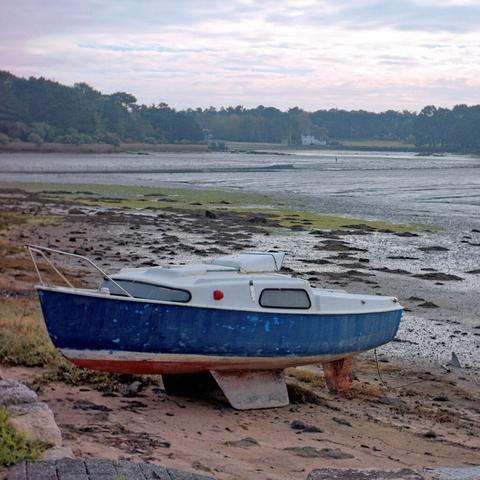 Photo de la mer a marrée basse. Au premier plan un bateau hors de l’eau, au loin de la forêt avec une maison, entre ces deux plans, des flaques d’eau.