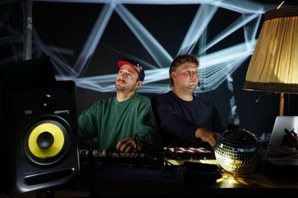 2 Personen hinter einem Technikpult (Lautsprecher, Mixer, Lampe, Diskokugel) im Hintergrund Lichtshow - Technostress