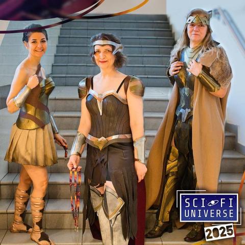 Tre donne in costume, che sembrano essere personaggi di fumetti o film fantasy, posano su una scala.  La donna a sinistra indossa un costume dorato e beige, simile a quello di Wonder Woman. La donna al centro ha un costume marrone e metallizzato, anch'esso simile a quello di Wonder Woman, ma con un design più scuro e dettagli più elaborati. La donna a destra indossa un costume più scuro con spallacci e un mantello color cammello, con un aspetto più guerriero.  Tutte e tre le donne hanno bracciali e protezioni metalliche. Dietro di loro, una scritta indica "Sci-Fi Universe 2025".
