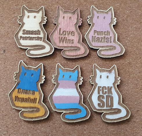 Photo of six lasercut cat magnets. five with captions "Smash patriarchy", "Love Wins", "Punch Nazis!", "Slava Ukraini" (in cyrilic), "FCKSD". One without text painted in a trans flag.