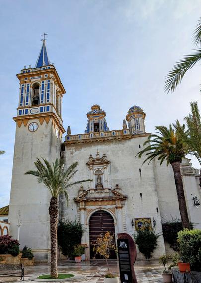 A church in a plaza. The church has white walls but the top of the pointed bellflower is a vivid blue. The sky is clear and blue and there are a few tall palm trees in front of the church.
