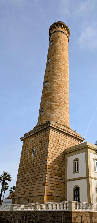 A tall, light brown bricked lighthouse. The Faro de Chipiona is the tallest in Spain, the third tallest in Europe and the fifth tallest in the world. The sky is blue and clear.