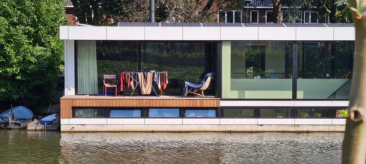 Primerily pink and salmon colored clothes drying in the sun on a clothes rack flanked by two chairs on the small terrace of a rectangular, modern design houseboat with large green tinted glass side panels.