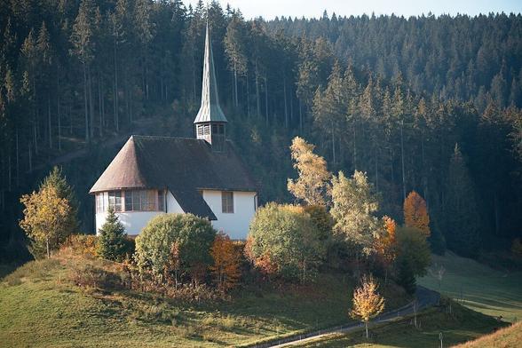 The image shows a small chapel with a steep spire located on a grassy hill. It is surrounded by trees with autumn foliage, and a dense forest forms the background. A narrow path runs near the hill, leading away from the structure.