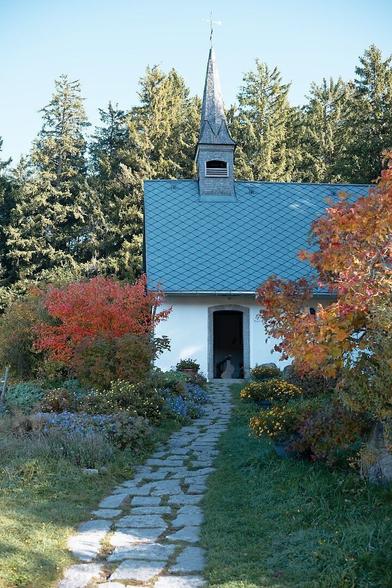The image depicts a small chapel with a steep roof and a spire featuring a weather vane. A stone path leads to the open entrance, flanked by colorful plants and shrubs, including red and yellow foliage. The background consists of tall evergreen trees.