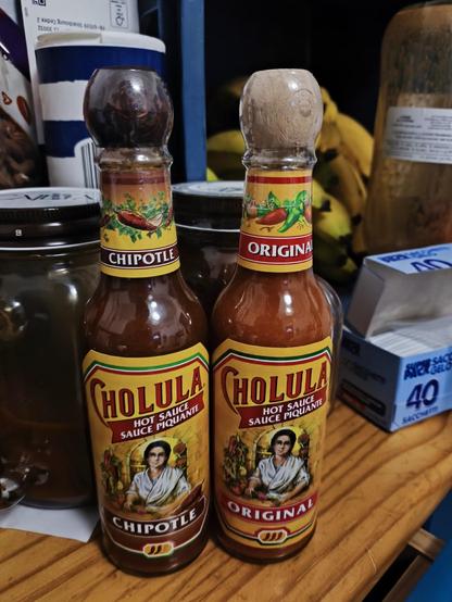 La imagen muestra dos botellas de salsa picante Cholula, colocadas sobre una superficie de madera. Ambas botellas tienen el característico diseño de la marca con etiquetas amarillas y rojas, y la ilustración de una mujer con un vestido blanco rodeada de ingredientes frescos como chiles y jitomates.

La botella de la izquierda es de sabor Chipotle y tiene una tapa de madera oscura brillante. En su etiqueta, se distingue un chile chipotle ilustrado junto a otros ingredientes. La botella de la derecha es la versión Original, con una tapa de madera más clara, casi beige. La etiqueta incluye un diseño similar, con chiles rojos y verdes destacados.

En el fondo, se observan otros objetos de cocina, como un tarro de vidrio, un paquete de bananas, un recipiente azul y blanco, y una caja de azúcar en sobres con la leyenda "40 Sachets." Estos elementos sugieren que la foto fue tomada en una cocina o despensa bien abastecida.