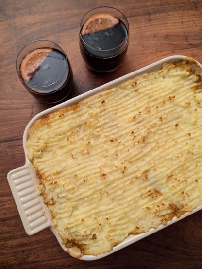 A rectangular photo of a potato-topped pie in a rectangular dish and two drinks in glasses, all sitting on a dark wooden background. The potato topping is covered in ridges, the high-points of which are golden. The drinks are dark red in colour, and each have a lemon wedge in them.