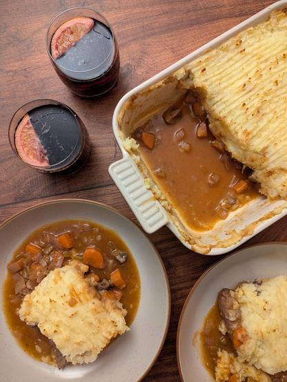 A rectangular photo of a potato-topped pie in a rectangular dish, two drinks in glasses, and two bowls with portions of the pie in them, all sitting on a dark wooden background. The top of the pie is covered in ridges, the tops of which are golden. A rectangle of the pie has been taken from its dish to put in the bowls, exposing a brown gravy and pieces of carrot, celery and onion inside. In the bowls there is the same mixture, and some sausages can be seen poking through. The drinks are dark red in colour, and each have a lemon wedge in them.