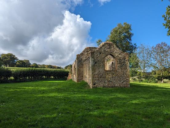 A ruined church built mostly of flint stones stands in a grass churchyard bound on the left by a low hedge and on the right by small trees and shrubs with a larger tree seen behind the ruins. The church is seen from the sanctuary end which has a large arched window space in the end wall. The sky on the right is clear and bright blue while on the left it is full of fluffy grey and white clouds.