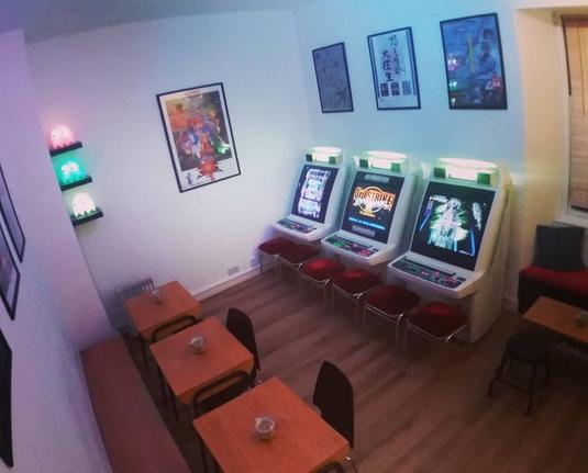 The back room at Konbo Arcade Cafe, with light-up Pac-Man ghosts stacked in the corner, arcade poster art on the walls, 3 Japanese arcade cabinets (candy cabs, all Astro City), stools, plus tables & chairs.