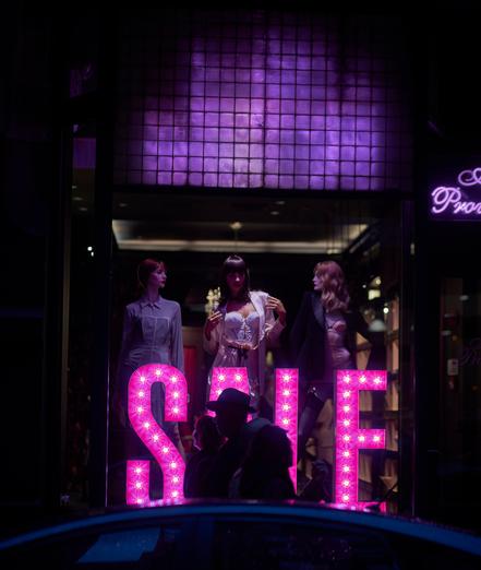 A well dressed family walks in front of a magenta SALE sign in a store window one evening, ignoring the mannequins posed in the window.