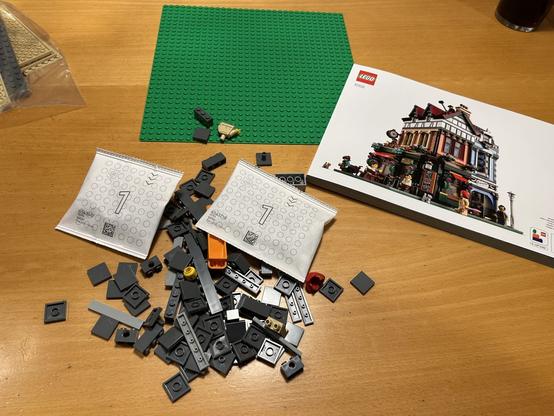 A green baseplate, instruction manual, two small paper bags with ‘1’ printed on them, and a loose pile of LEGO elements, all sitting on a wooden table, ready to begin building
