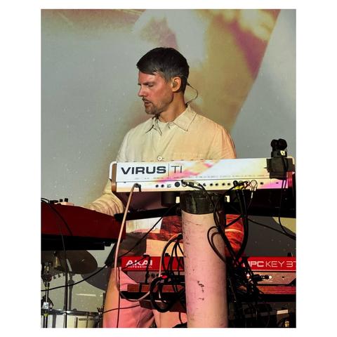 A concert photo of Tycho, capture mid-performance. Scott Hansen, dressed in all white pants and a button up shirt, is performing behind a stand holding various synthesizers and keyboards, some of which are a Virus TI and a AKAI MPC Key 37. Cables are pouring out of his set up as warm beige, washed out visuals are projected onto and behind him.