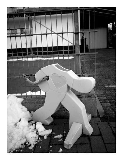 Black and white photo of a reflecting child figure to slow traffic. The figure shows a big heart, but the head is missing. It is also chained to a metal fence.