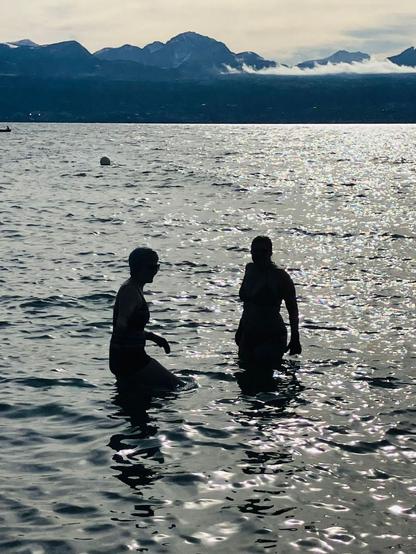 Silhouettes de 2 femmes en contre jour qui se baignent dans le lac avec les montagnes derrière.