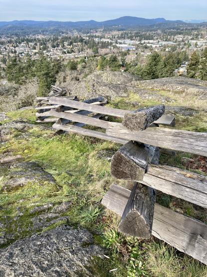 A smooth rocky crown of hill, lots of grass and moss showing. Marking the path is an old split log rail fence. You can see houses below and a distant mountain range.