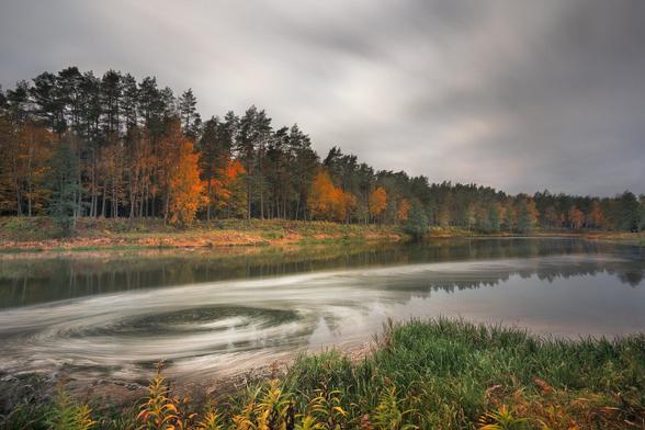 Long exposure of the river in the woods on a cloudy day. Trees are all autumn colors and reflected on a smooth long exposed water. Also big swirls of water bubble trails are seen on the river foreground