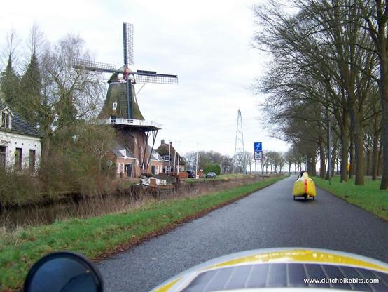 Are we in the Netherlands ? The photo shows a cyclepath alongside a canal, with velomobiles on the cyclepath and a traditional windmill on the other side of the canal. Looks very Dutch.