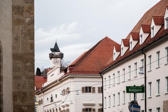 View of the clocktower of Graz. On the left side there is a part of a church. Under the Uhrturm there are old houses.