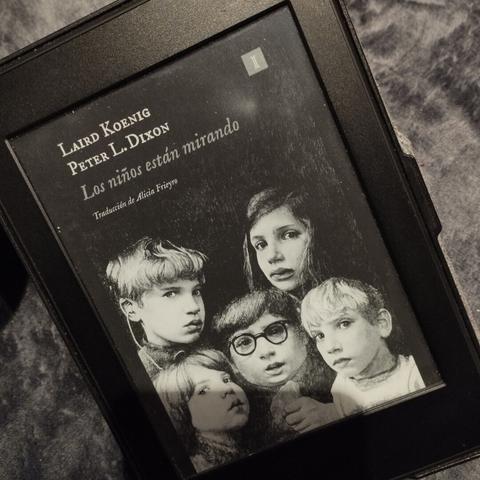 Un Kindle negro sobre fondo gris mostrando la portada en blanco y negro del libro «Los niños están mirando», de Laird Koening. Aparecen tres niños y dos niñas mirando hacia el espectador.