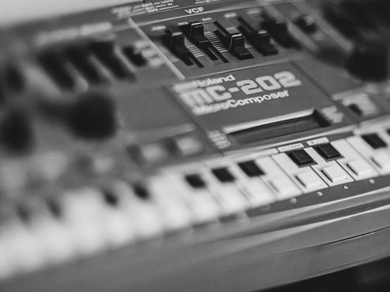 A close-up, black and white photo of a Roland MC-202 MicroComposer keyboard. The photo shows various knobs, switches, and the name "Roland MC-202 MicroComposer" in focus.