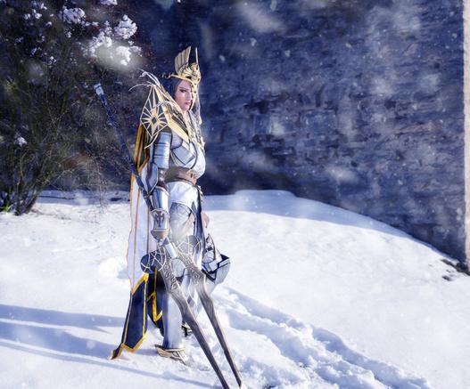 This is a photo of a cosplayer: A person wearing armor is standing in the snow. She wears huge pauldrons, a cape, a headpiece in the shape of an eagle and half of her face is covered by a golden mask. In her hand she is holding a forked spear with two blades.