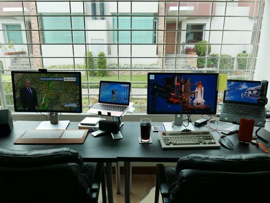 Two desks, side by side, located in a window on the ground floor. Outside the window is bright daylight, with two well-manicured Latin American townhomes visible across the way. The desks each have a laptop on a stand; a keyboard; a mouse/trackpad; and a monitor connected to them. The desk on the left: a pink Macbook Retina, connected to a 24" monitor. It has a white wireless Mac keyboard, and matching trackpad. The monitor has local news broadcast on it. The right desk: a ThinkPad running OpenBSD, connected to its own monitor. The monitor displays the cwm window manager, and xclock. The wallpaper is visible, showing a space shuttle about to launch. The keyboard is a white/beige Model M SSK, white. There is a trackball on this desk. The desks are adjacent to one another, each with matching chairs. There is a fur rug underneath both desks where one's feet would be.