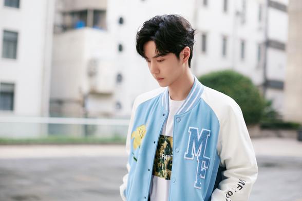 Wang Yibo poses for a promo photo while wearing a pale blue varsity style jacket with a yellow lily on one side, and the letters MF on the other side. He's looking down, and has his hands in the pockets of the jacket.