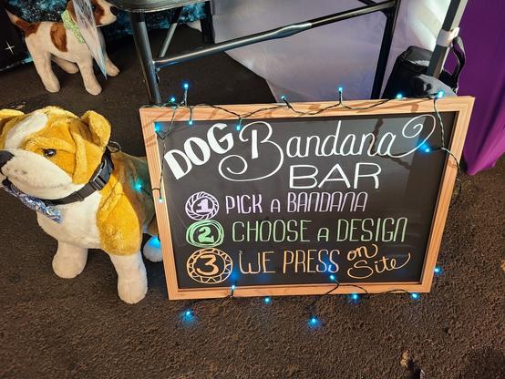 Chalkboard sign that says dog bandana bar 1 pick a bandana 2 choose a design 3 we press on site. a stuffed bulldog is sitting next to the sign
