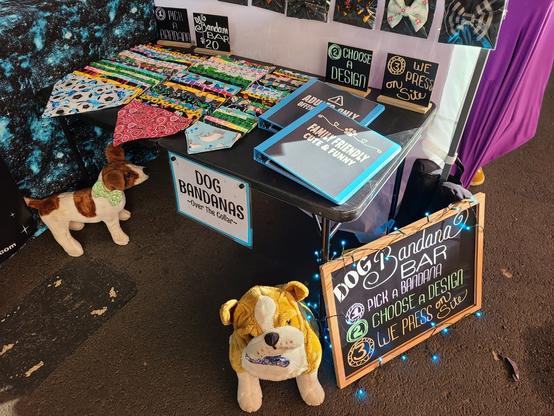 Table with the bandanas and notebooks and the sign in front of the table. 2 stuffed dogs sitting and standing nearby