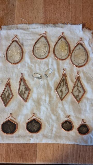 Some glass pairs of earrings with their bails detached above them. All contours and bails look like copper. After they're soldered they will look silvery. Between the glass planes are lunaria discs, plants roots, and skeleton hydrangea petals. The latter have a black background so they stand out more on the skin.