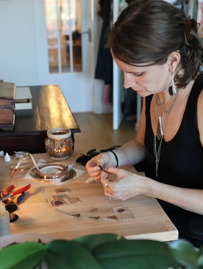 A craftswoman works at her desk, with glass planes, copper tape and wire containing plant parts. She's wearing the jewelry she makes: stained glass earrings with silvery contour and dragonfly wings inside, and a long necklace with intricate cascades of chains surrounding a fern in a stained glass pendant.