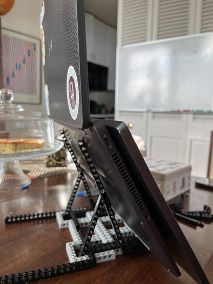 A laptop is propped up at an angle on a stand made of LEGO. The background features a faint glimpse of a kitchen, a whiteboard, and a cake under a glass dome on a wooden table. Stickers are visible on the laptop's cover
