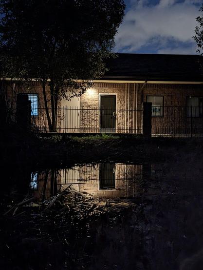 A nighttime scene of a small building softly illuminated by warm lights, with a dark, quiet pond in the foreground. The pond creates a perfect reflection of the building, resembling an inverted, dreamlike world. Shadows from surrounding trees frame the image, adding depth and a sense of mystery. The stillness of the water contrasts with the textured building, evoking a tranquil and otherworldly atmosphere.