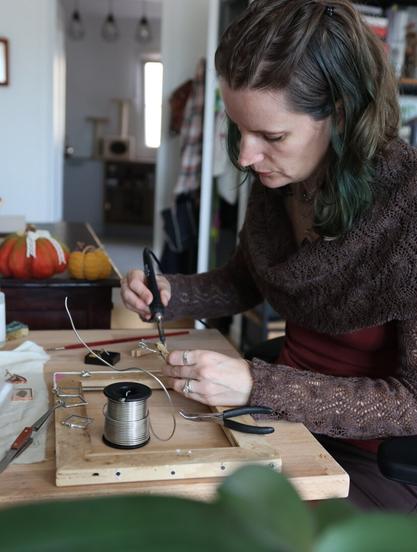 A craftswoman works at her desk, she's soldering stained glass pendants with an iron. Her hair is half dark forest green, half dark blonde.