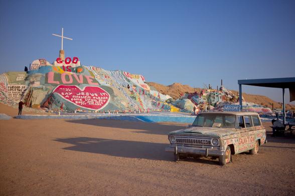 Wide shot of salvation mountain. Paint covered rock hill with "Got is Love" written across the top. An old abandoned car is also covered in paint and decorations.