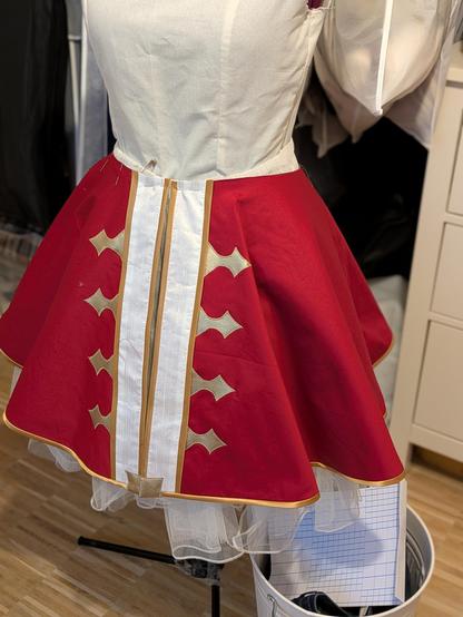 3/4 circle skirt in red with golden trim and applique and open center front, white appliques to the center. It's put on a dressform with a petticoat