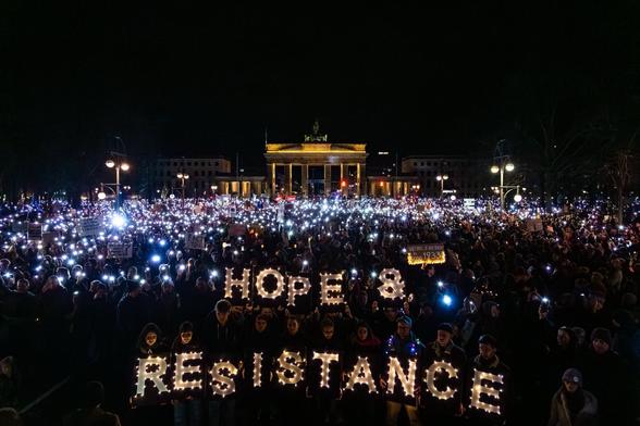 A powerful nighttime image capturing a crowd of approximately 100,000 people in Berlin, gathered in front of the illuminated Brandenburg Gate. Participants hold glowing signs spelling "HOPE & RESISTANCE," with thousands of lights from phones and lanterns illuminating the scene. Protesters carry banners and signs, including one warning against the normalization of far-right ideologies, referencing historical parallels like 1933. The image symbolizes solidarity and resistance against fascism in Germany and across Europe.