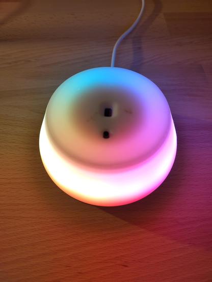 Ein Bild der Glowlight-Lampe im eingeschalteten Zustand. Die Lampe strahlt ein sanftes, buntes Licht in verschiedenen Farben (RGB) aus und steht auf einer Holzoberfläche. Ein Stromkabel ist im Hintergrund zu sehen.