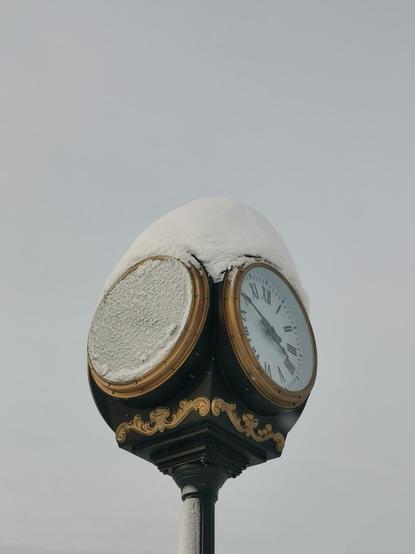 This image showcases a vintage-style outdoor clock, elegantly adorned with gold detailing and Roman numerals, partially covered in a thick layer of snow. The clock stands against a pale, overcast winter sky, emphasizing the serene and chilly atmosphere. The snow accentuates the ornate design of the clock, adding a sense of stillness and timelessness to the scene.