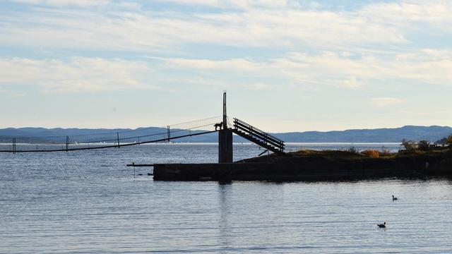A photo of a small suspension footbridge across water. It leads to a gate and stairs going down to the ground. There is a dog standing on the bridge at the gate. There are hills in the distance. The sky is a hazy blue with white clouds.