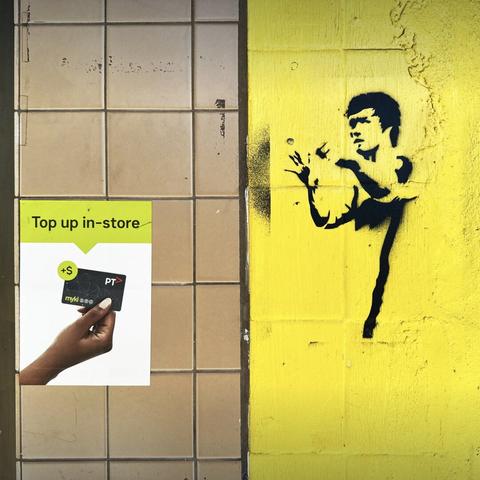 A close-up of a wall, on the left, a sticker on a tiled wall advertising “top up in store” with a hand holding a public transport card; on the right, a yellow wall with a black stencil of someone (Bruce Lee?) making some hand gestures in the direction of the sticker