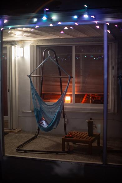 a blue hammock chair cradling a laptop, in a porch under darkness with opalescent string lights around the periphery, next to a small square ikea table with a speaker, a phone, and a glass of red beverage on it
