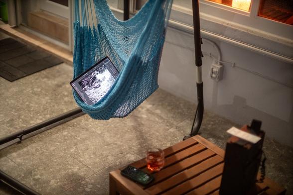a blue hammock chair cradling a laptop with a black and white picture on the screen, next to a small square ikea table with a speaker, a phone, and a glass of red beverage on it