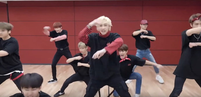 Stray Kids in a dance practice studio. Bang Chan is in the centre. Seungmin is on one knee behind him, reaching towards Chan's butt with an expression of open-mouthed glee.