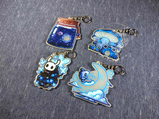 Blue keychains featuring some frogs sitting under mushrooms, mola in a jar, mola in the clouds on a moon, and the knight from Hollow Knight sitting on a bench.