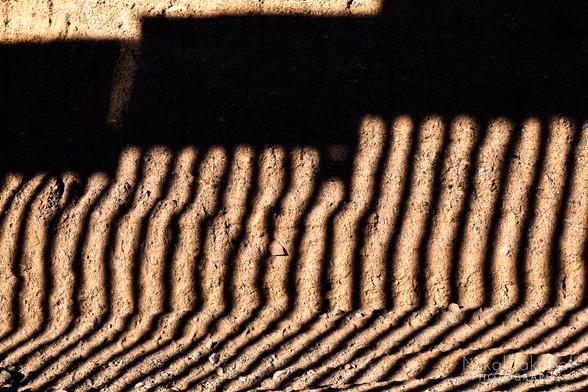 A dirt cutbank alongside a road. The bank is striped with vertical shadows like bars of a cell, cast by the low afternoon sun shining through the U.S.-Mexico border fence. Across the top of the striped shadows is a solid dark horizontal shadow cast by the solid steel plate atop the fence.