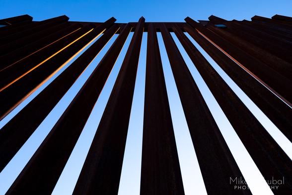 A steep view looking directly upwards from the base to the top of the U.S.-Mexico border fence. The angular bars slightly converge at they approach the steel plate on top of the fence. Because the "bars" of the fence are square tubes turned 45°, the sun strikes the angled faces of some of the bars, lighting up the rusty orange of the exposed iron. The side toward the viewer is in shadow, creating a striking contrast between the orange sunlit faces of the bars and the bright blue sky behind the bars.