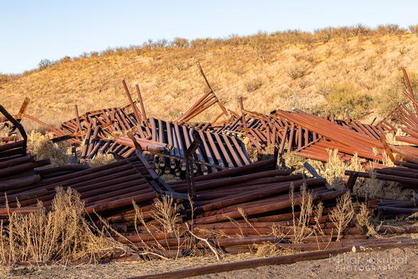 An open field with a dry hill covered in scrubby brush behind it. In the field is a jumbled pile of sections of old border fence. Each section is made of concrete-filled steel tubes about 20' long, torn out in big chunks and thrown hodgepodge in a heap.
