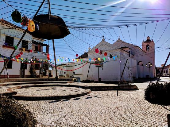 A festive scene in a small town square. Colorful paper lanterns, bunting, and a black umbrella hang from a large net-like structure overhead, creating a cheerful atmosphere. A whitewashed church stands in the background.